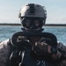 A military combat diver is photographed in close-up wearing full tactical diving gear, including a rebreather mask with corrugated hoses, a tan ballistic helmet with a night-vision mount, and desert camouflage uniform. The subject appears to be in or near open water, with a coastline visible in the background. The image captures a special operations or combat diving context based on the specialized equipment visible.