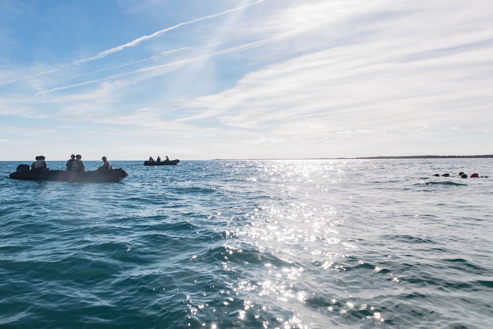 The image shows an open ocean scene with two black inflatable rubber boats (IRBs/RHIBs) carrying multiple personnel, likely military or special operations based on their equipment and posture. To the right of frame, several individuals are visible in the water, appearing to swim or float near the surface. The scene is backlit by sunlight reflecting off calm-to-moderate seas under a partly cloudy sky.