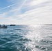 The image shows an open ocean scene with two black inflatable rubber boats (IRBs/RHIBs) carrying multiple personnel, likely military or special operations based on their equipment and posture. To the right of frame, several individuals are visible in the water, appearing to swim or float near the surface. The scene is backlit by sunlight reflecting off calm-to-moderate seas under a partly cloudy sky.