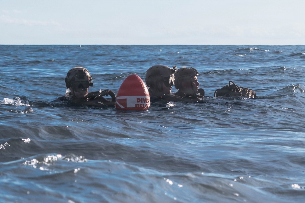 Three military divers are visible at the surface of open ocean water, equipped with tactical diving gear including helmets and rebreather/scuba equipment. A red and white dive marker buoy with partially visible text reading 'DIVER BELOW' is positioned between them. The image appears to document a military combat diving or special operations training exercise.