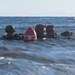 Three military divers are visible at the surface of open ocean water, equipped with tactical diving gear including helmets and rebreather/scuba equipment. A red and white dive marker buoy with partially visible text reading 'DIVER BELOW' is positioned between them. The image appears to document a military combat diving or special operations training exercise.