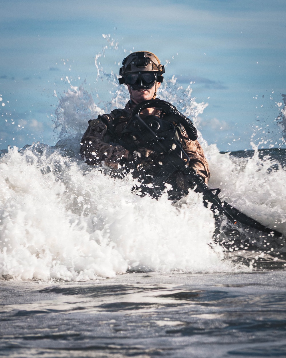 A heavily equipped military combat diver or special operations soldier is emerging from ocean surf onto a beach. The individual is wearing full tactical gear including a helmet, diving rebreather apparatus, and appears to be carrying a weapon. The image captures a dynamic amphibious insertion or beach landing exercise.