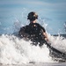 A heavily equipped military combat diver or special operations soldier is emerging from ocean surf onto a beach. The individual is wearing full tactical gear including a helmet, diving rebreather apparatus, and appears to be carrying a weapon. The image captures a dynamic amphibious insertion or beach landing exercise.