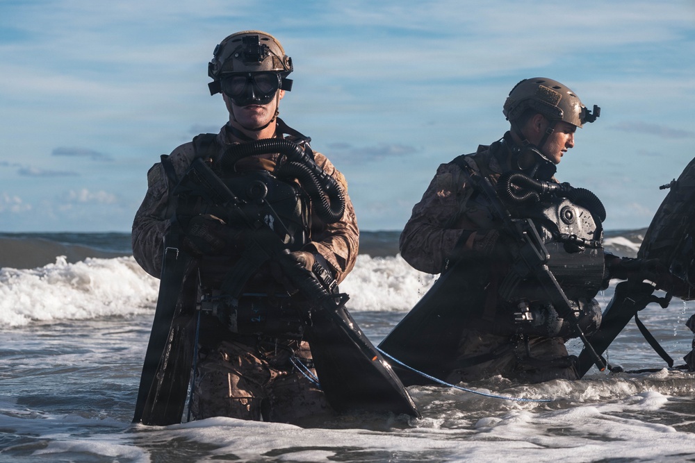 Two heavily equipped military personnel are emerging from ocean surf onto a beach, carrying weapons and wearing full combat gear including helmets with night-vision device mounts and closed-circuit rebreather diving apparatus. The image appears to depict a combat swimmer or amphibious insertion exercise, consistent with special operations forces training. The scene is captured in sharp daylight conditions with breaking waves visible in the background.