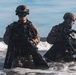 Two heavily equipped military personnel are emerging from ocean surf onto a beach, carrying weapons and wearing full combat gear including helmets with night-vision device mounts and closed-circuit rebreather diving apparatus. The image appears to depict a combat swimmer or amphibious insertion exercise, consistent with special operations forces training. The scene is captured in sharp daylight conditions with breaking waves visible in the background.