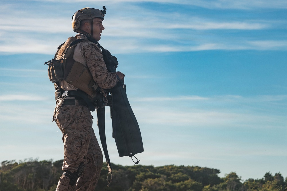 A military service member in full combat gear stands in a profile view against a partly cloudy blue sky. The individual is wearing desert MARPAT camouflage uniform consistent with U.S. Marine Corps attire, equipped with a tactical vest, helmet with mounted device, and backpack. The soldier appears to be holding what looks like diving fins, suggesting a maritime or amphibious training context.