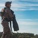 A military service member in full combat gear stands in a profile view against a partly cloudy blue sky. The individual is wearing desert MARPAT camouflage uniform consistent with U.S. Marine Corps attire, equipped with a tactical vest, helmet with mounted device, and backpack. The soldier appears to be holding what looks like diving fins, suggesting a maritime or amphibious training context.