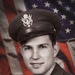A black-and-white formal military portrait photograph of a young male serviceman, composited against a colorized American flag background. The subject wears a U.S. military service cap with an eagle insignia and a dark uniform with tie. The image appears to be a mid-20th century official or semi-official military portrait, likely from the World War II or Korean War era.