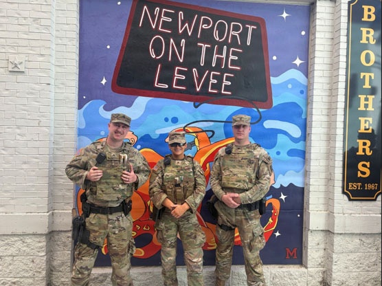 Three military personnel in camouflage uniforms and tactical gear are posing in front of a colorful mural at Newport on the Levee, a entertainment district in Newport, Kentucky. They appear to be on duty, wearing body armor and carrying equipment. The photo appears to be a casual group photo taken at a recognizable landmark.