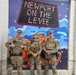 Three military personnel in camouflage uniforms and tactical gear are posing in front of a colorful mural at Newport on the Levee, a entertainment district in Newport, Kentucky. They appear to be on duty, wearing body armor and carrying equipment. The photo appears to be a casual group photo taken at a recognizable landmark.
