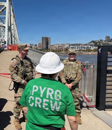 Three individuals are standing on a bridge deck near a river, with a city skyline visible in the background. Two individuals are wearing military camouflage uniforms with tactical vests, while a third person faces away from the camera wearing a green 'Pyro Crew 2020' t-shirt and a white hard hat. The setting suggests a coordinated event involving pyrotechnics, likely a fireworks show setup with military security presence.