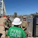 Three individuals are standing on a bridge deck near a river, with a city skyline visible in the background. Two individuals are wearing military camouflage uniforms with tactical vests, while a third person faces away from the camera wearing a green 'Pyro Crew 2020' t-shirt and a white hard hat. The setting suggests a coordinated event involving pyrotechnics, likely a fireworks show setup with military security presence.
