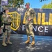 Three military personnel in U.S. Army OCP (Operational Camouflage Pattern) uniforms are gathered outdoors near a banner reading 'HILLS FENCE CO.' A civilian woman in a blue dress and a man in a black shirt are also present, suggesting a community or public event. The soldiers appear to be engaged in conversation, possibly at a promotional or recruitment event.