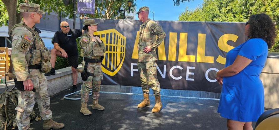Three military personnel in U.S. Army OCP (Operational Camouflage Pattern) uniforms are gathered outdoors near a banner reading 'HILLS FENCE CO.' A civilian woman in a blue dress and a man in a black shirt are also present, suggesting a community or public event. The soldiers appear to be engaged in conversation, possibly at a promotional or recruitment event.