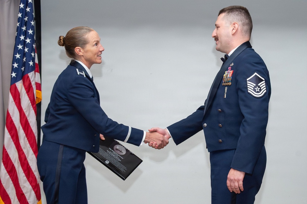 Two United States Air Force personnel in dress blue uniforms are shaking hands during what appears to be a formal military ceremony. The female officer on the left is holding a plaque or award, suggesting this is a recognition or commendation event. An American flag is visible in the background to the left.