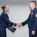 Two United States Air Force personnel in dress blue uniforms are shaking hands during what appears to be a formal military ceremony. The female officer on the left is holding a plaque or award, suggesting this is a recognition or commendation event. An American flag is visible in the background to the left.