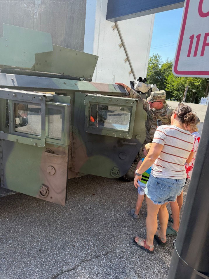 A uniformed military soldier is showing a military armored vehicle to civilians, including a woman and at least one child, in what appears to be a public display or recruitment event. The vehicle is olive drab green and appears to be a tracked armored personnel carrier or similar military vehicle with its door open. The setting appears to be a parking lot or fairground area on a sunny day.
