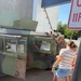 A uniformed military soldier is showing a military armored vehicle to civilians, including a woman and at least one child, in what appears to be a public display or recruitment event. The vehicle is olive drab green and appears to be a tracked armored personnel carrier or similar military vehicle with its door open. The setting appears to be a parking lot or fairground area on a sunny day.