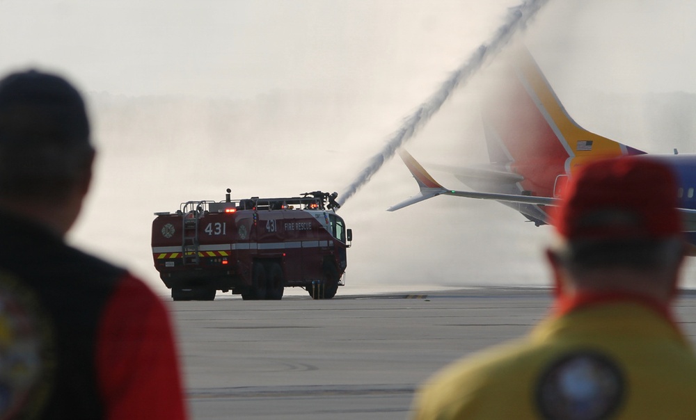 An airport fire rescue vehicle (unit 431) is spraying water onto a Southwest Airlines aircraft on an airport tarmac. Two personnel in the foreground observe the operation, which appears to be either an emergency response or a ceremonial water cannon salute. The scene takes place on an open tarmac with hazy atmospheric conditions.
