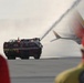 An airport fire rescue vehicle (unit 431) is spraying water onto a Southwest Airlines aircraft on an airport tarmac. Two personnel in the foreground observe the operation, which appears to be either an emergency response or a ceremonial water cannon salute. The scene takes place on an open tarmac with hazy atmospheric conditions.