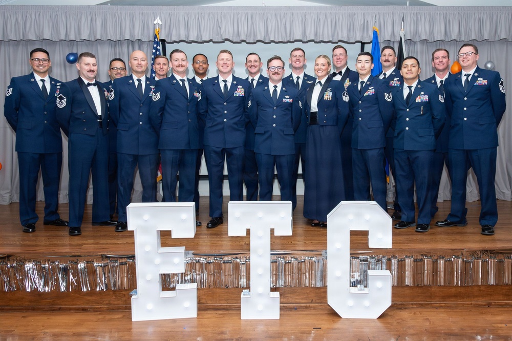 A group of approximately 18-19 U.S. Air Force personnel in dress blue uniforms pose for a formal group photograph on a stage. In the foreground, large illuminated marquee letters spell 'ETC', likely representing a specific course, unit, or event. The setting appears to be a formal military banquet or graduation ceremony.