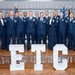 A group of approximately 18-19 U.S. Air Force personnel in dress blue uniforms pose for a formal group photograph on a stage. In the foreground, large illuminated marquee letters spell 'ETC', likely representing a specific course, unit, or event. The setting appears to be a formal military banquet or graduation ceremony.