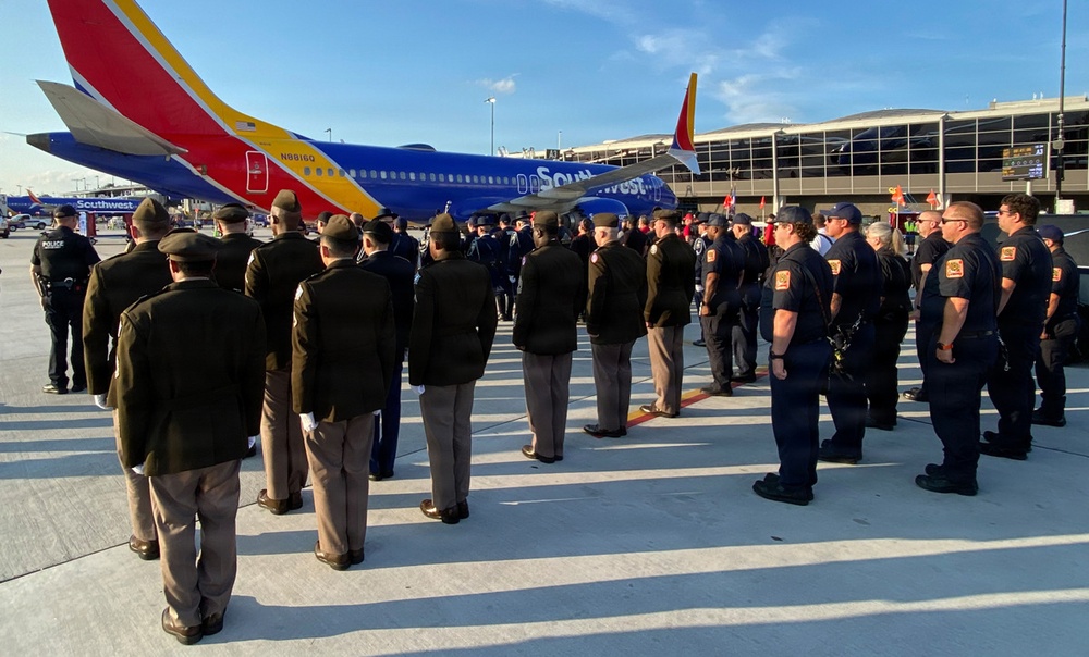A formal ceremonial gathering is taking place on an airport tarmac in front of Southwest Airlines aircraft. Multiple uniformed personnel — including what appear to be law enforcement officers, military members, and security staff — are assembled in organized formations. The scene suggests a dignitary arrival, fallen officer tribute, or official honors ceremony.