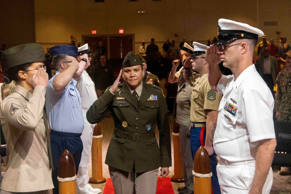 A military ceremony is taking place in what appears to be a large indoor venue, with service members from multiple branches of the U.S. Armed Forces rendering salutes. A central figure, a female U.S. Army officer in dress greens with a garrison cap and multiple ribbons/decorations, is walking through a corridor formed by saluting personnel. The event appears to be a formal military honors ceremony, possibly a promotion, retirement, or change of command.