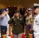A military ceremony is taking place in what appears to be a large indoor venue, with service members from multiple branches of the U.S. Armed Forces rendering salutes. A central figure, a female U.S. Army officer in dress greens with a garrison cap and multiple ribbons/decorations, is walking through a corridor formed by saluting personnel. The event appears to be a formal military honors ceremony, possibly a promotion, retirement, or change of command.