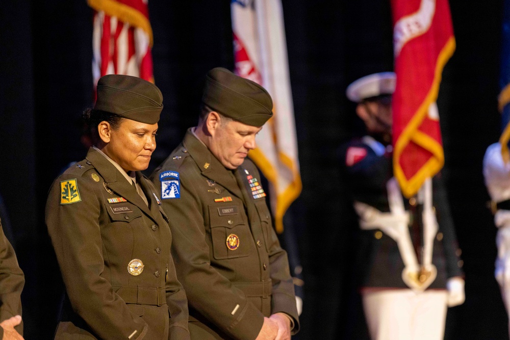 Two U.S. Army officers in Army Green Service Uniforms stand with heads bowed during what appears to be a formal military ceremony. The female officer on the left wears a brigadier general star and name tape reading 'McDENEGAL,' while the male officer on the right displays Airborne and 18th Airborne Corps patches with name tape reading 'EBBERT.' Multiple military flags and a color guard are visible in the background.