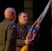 A military ceremony is taking place in which a soldier identified by name tape as 'EBBERT' is handling a unit guidon or organizational flag with campaign streamers. The flag appears to bear the text 'USNORTHCOM' or similar command designation, suggesting this is a change of command or activation ceremony. Three military personnel in Army service uniforms are visible, with one wearing a visible airborne patch.