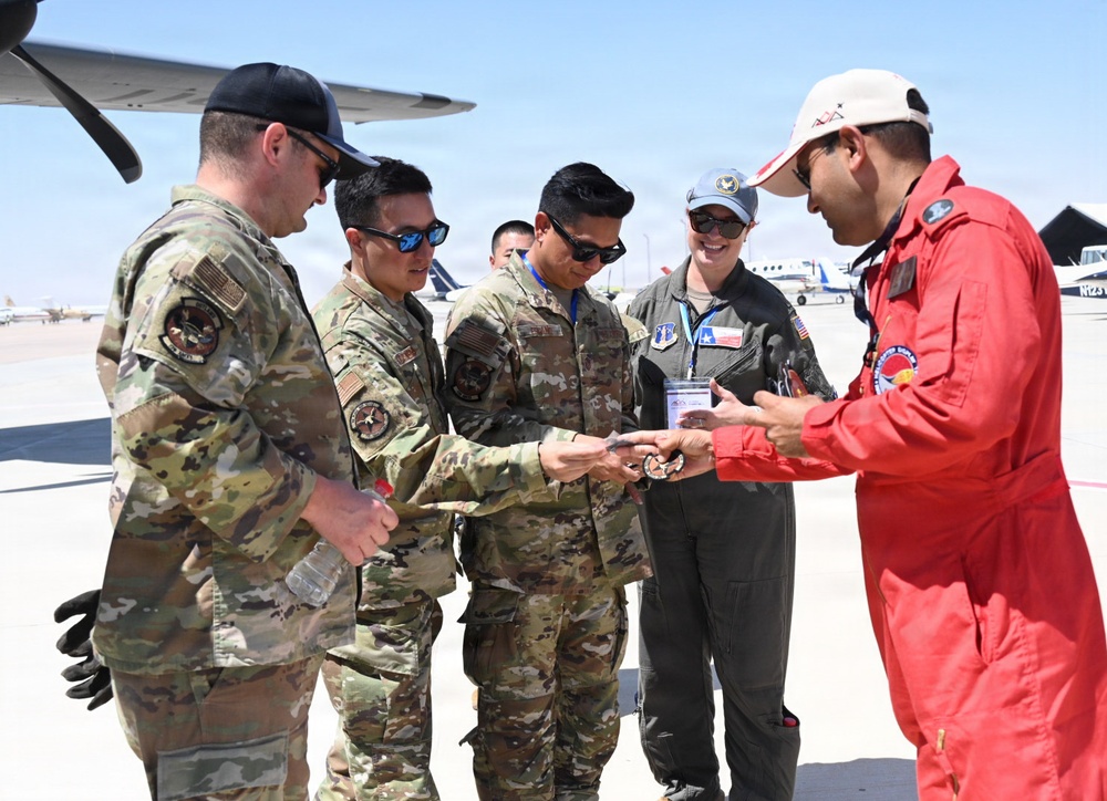 A group of military personnel in US Army/Air Force camouflage uniforms are gathered on an airfield tarmac with an individual wearing a red flight suit. They appear to be exchanging or examining challenge coins or small items, a common military tradition. Aircraft and airport infrastructure are visible in the background.