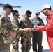A group of military personnel in US Army/Air Force camouflage uniforms are gathered on an airfield tarmac with an individual wearing a red flight suit. They appear to be exchanging or examining challenge coins or small items, a common military tradition. Aircraft and airport infrastructure are visible in the background.