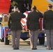 Three uniformed military personnel are standing at attention and saluting a flag-draped casket on a baggage cart at an airport tarmac. The casket is draped with what appears to be an American flag, indicating a dignified transfer ceremony. The aircraft visible in the background appears to be a Southwest Airlines commercial plane.