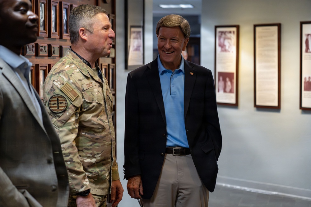 Three individuals are standing in what appears to be a military facility hallway or corridor, with framed photographs and plaques displayed on the walls. A military service member in Army Combat Uniform (ACU/OCP camouflage) is conversing with a civilian man wearing a blue polo shirt and dark blazer. The setting suggests an official visit or tour of a military installation.