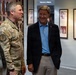 Three individuals are standing in what appears to be a military facility hallway or corridor, with framed photographs and plaques displayed on the walls. A military service member in Army Combat Uniform (ACU/OCP camouflage) is conversing with a civilian man wearing a blue polo shirt and dark blazer. The setting suggests an official visit or tour of a military installation.