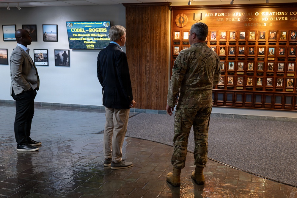 Three individuals are standing in a hallway at Air Force Special Operations Command (AFSOC), viewing a memorial wall displaying Air Force Cross and Silver Star recipients. A digital sign identifies the visit as 'CODEL-ROGERS' welcoming The Honorable Mike Rogers, Chairman of the House Armed Services Committee. One person is in military camouflage uniform while the other two are in civilian attire.