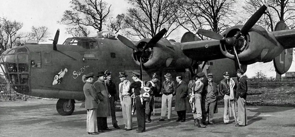 A black-and-white photograph showing a group of approximately 12-15 military personnel gathered in front of a large four-engine heavy bomber aircraft. The aircraft has nose art visible, appearing to read 'Suzy' along with a painted figure, and mission markers are visible below the cockpit. The setting appears to be an airfield with bare trees in the background, consistent with a WWII-era scene.