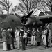 A black-and-white photograph showing a group of approximately 12-15 military personnel gathered in front of a large four-engine heavy bomber aircraft. The aircraft has nose art visible, appearing to read 'Suzy' along with a painted figure, and mission markers are visible below the cockpit. The setting appears to be an airfield with bare trees in the background, consistent with a WWII-era scene.