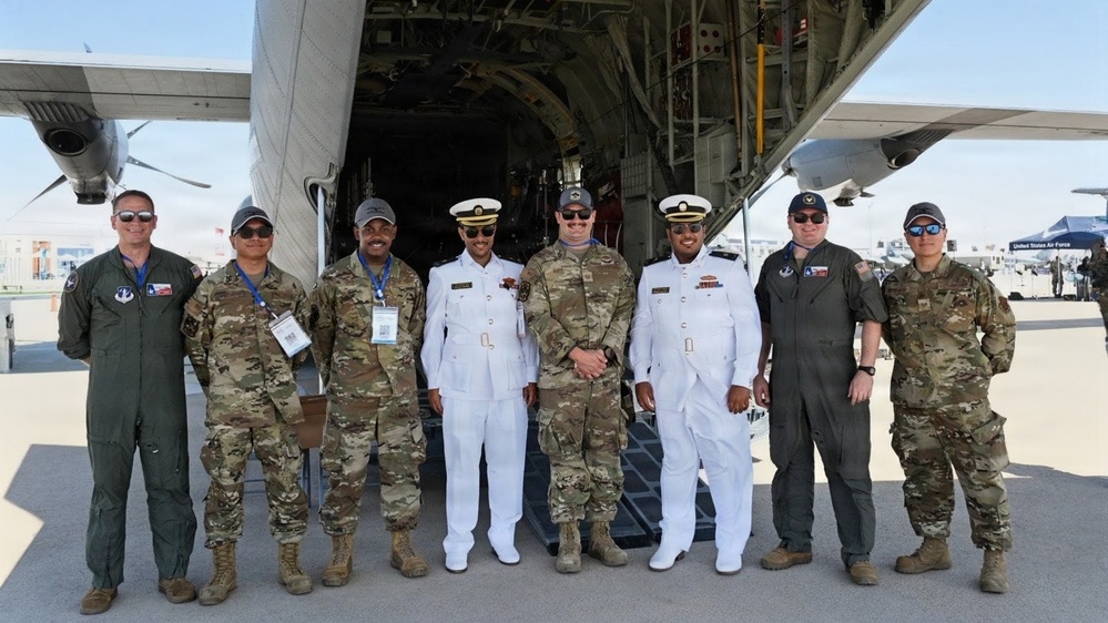 A group of eight military personnel pose for a photo in front of a large military transport aircraft with its rear cargo ramp open. The group includes personnel in U.S. Air Force flight suits and camouflage uniforms, as well as two individuals in white naval dress uniforms, suggesting a multinational or inter-service gathering. A 'United States Air Force' sign is visible in the background, indicating this appears to be an air show or official military event.