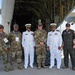 A group of eight military personnel pose for a photo in front of a large military transport aircraft with its rear cargo ramp open. The group includes personnel in U.S. Air Force flight suits and camouflage uniforms, as well as two individuals in white naval dress uniforms, suggesting a multinational or inter-service gathering. A 'United States Air Force' sign is visible in the background, indicating this appears to be an air show or official military event.
