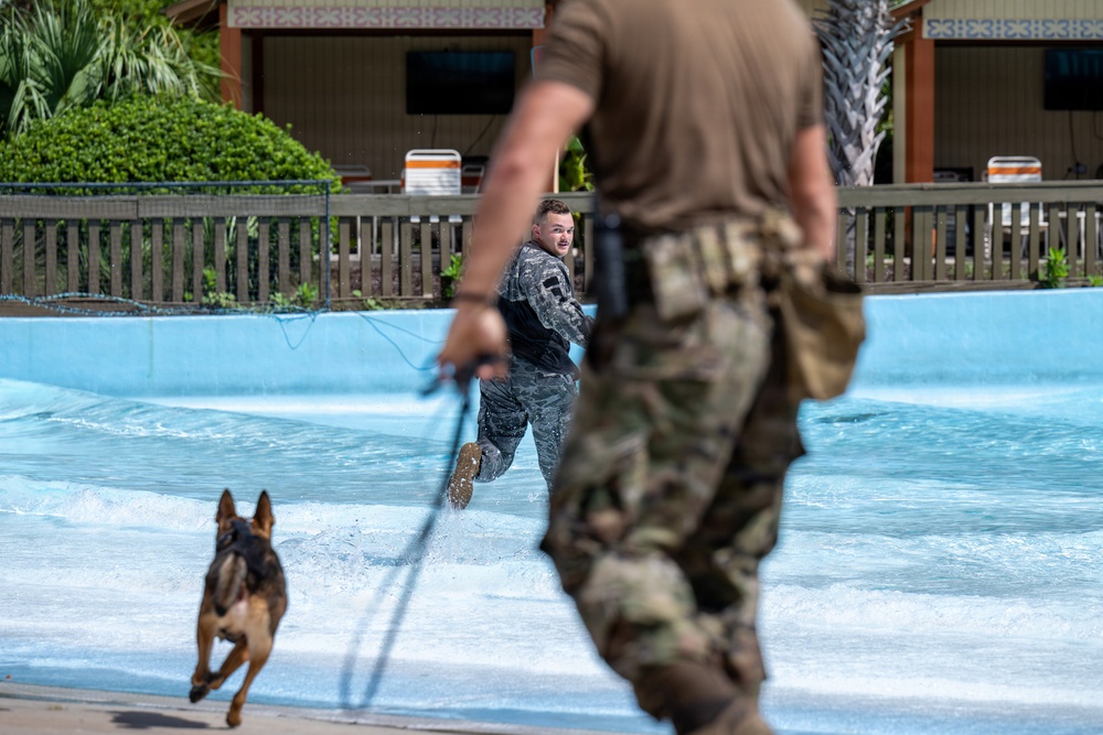 A military working dog (MWD) training exercise is taking place in what appears to be a wave pool or shallow water feature at a recreational facility. A person in military camouflage uniform is running through the water while a German Shepherd on a leash pursues or accompanies them. A second uniformed handler in the foreground holds the leash, overseeing the exercise.