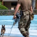 A military working dog (MWD) training exercise is taking place in what appears to be a wave pool or shallow water feature at a recreational facility. A person in military camouflage uniform is running through the water while a German Shepherd on a leash pursues or accompanies them. A second uniformed handler in the foreground holds the leash, overseeing the exercise.