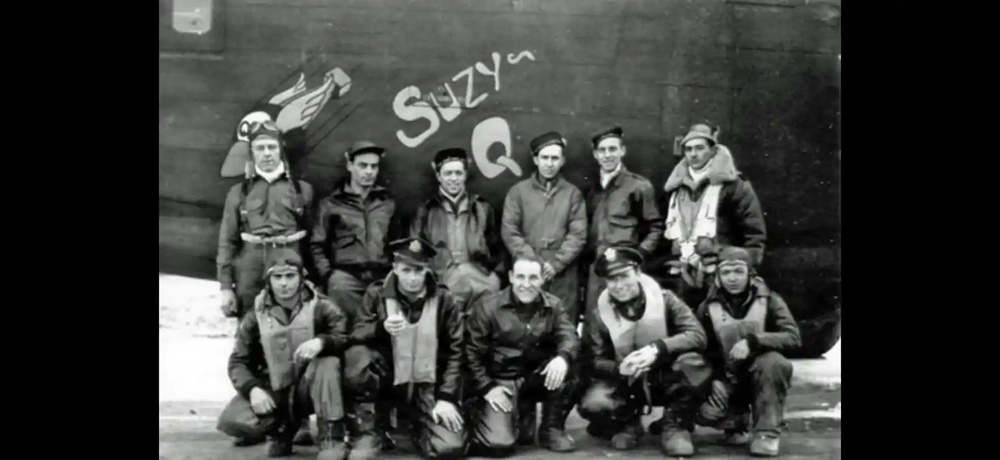 A black-and-white World War II era photograph showing a crew of approximately eleven airmen posed in front of a military aircraft named 'Suzy Q'. The men are wearing typical WWII-era flight gear including leather A-2 jackets, flight suits, and various headgear. The nose art 'Suzy Q' is clearly visible on the dark fuselage behind them.