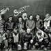 A black-and-white World War II era photograph showing a crew of approximately eleven airmen posed in front of a military aircraft named 'Suzy Q'. The men are wearing typical WWII-era flight gear including leather A-2 jackets, flight suits, and various headgear. The nose art 'Suzy Q' is clearly visible on the dark fuselage behind them.