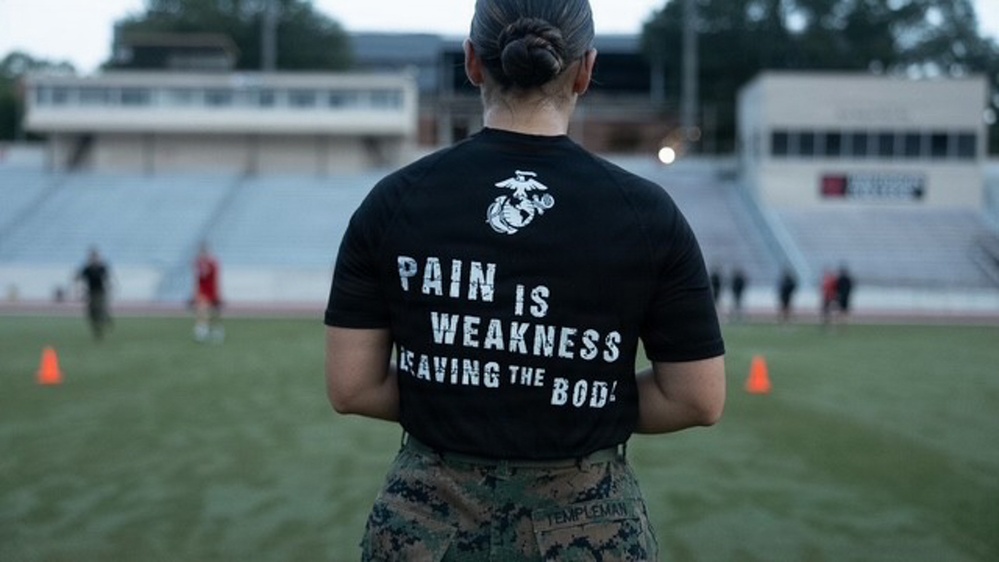 A person wearing a black U.S. Marine Corps t-shirt stands with their back to the camera on a sports field or track, overlooking what appears to be a physical training session. The shirt reads 'PAIN IS WEAKNESS LEAVING THE BODY' with the USMC Eagle, Globe, and Anchor emblem. The individual is wearing military camouflage (MARPAT) pants with a name tape reading 'TEMPLEMAN'.