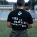A person wearing a black U.S. Marine Corps t-shirt stands with their back to the camera on a sports field or track, overlooking what appears to be a physical training session. The shirt reads 'PAIN IS WEAKNESS LEAVING THE BODY' with the USMC Eagle, Globe, and Anchor emblem. The individual is wearing military camouflage (MARPAT) pants with a name tape reading 'TEMPLEMAN'.