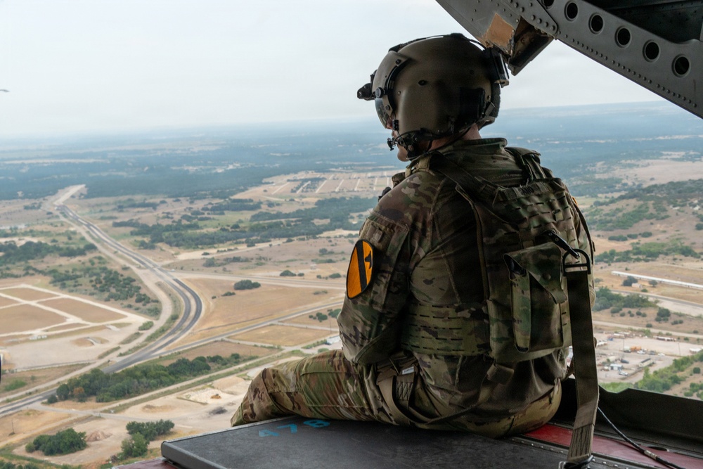 A U.S. Army soldier wearing MultiCam (OCP) camouflage and a flight helmet sits at the open door or ramp of a military helicopter, looking out over a vast aerial landscape. The soldier displays a 1st Cavalry Division patch (black and yellow shield) on the left shoulder. The terrain below appears to be semi-arid flatlands consistent with central Texas, likely near Fort Cavazos (formerly Fort Hood).