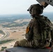A U.S. Army soldier wearing MultiCam (OCP) camouflage and a flight helmet sits at the open door or ramp of a military helicopter, looking out over a vast aerial landscape. The soldier displays a 1st Cavalry Division patch (black and yellow shield) on the left shoulder. The terrain below appears to be semi-arid flatlands consistent with central Texas, likely near Fort Cavazos (formerly Fort Hood).