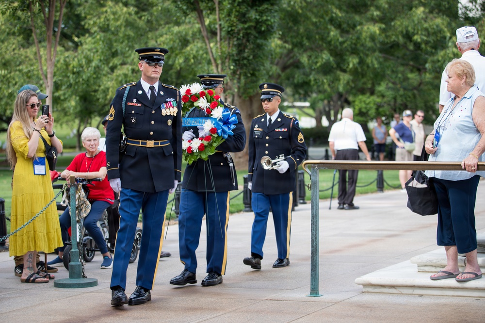 A military wreath-laying ceremony is taking place at what appears to be a national memorial or cemetery, likely Arlington National Cemetery. Uniformed U.S. Army soldiers in dress blues are carrying a red, white, and blue floral wreath while civilians and veterans observe from the sides. The formal procession suggests a commemorative or memorial event.